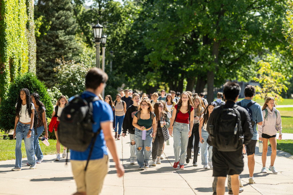 Several young men and women carrying backpacks walk up a tree-lined sidewalk.