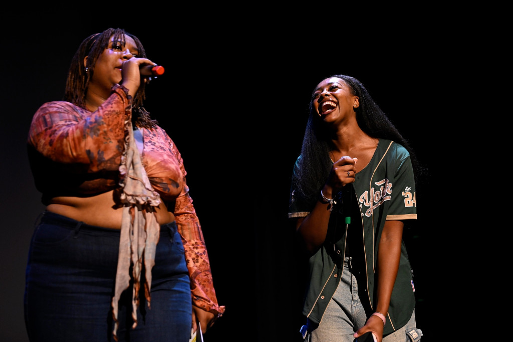 Two people on stage talk into microphones and laugh.