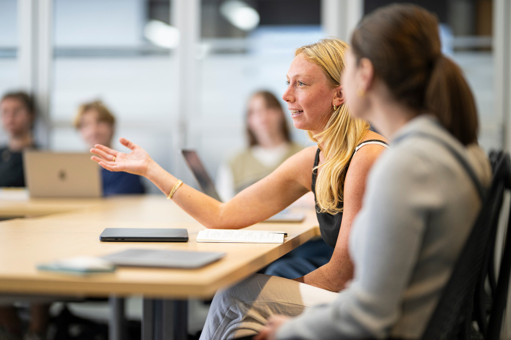 A woman gestures with her hands while speaking. She's seated at a table with other students.