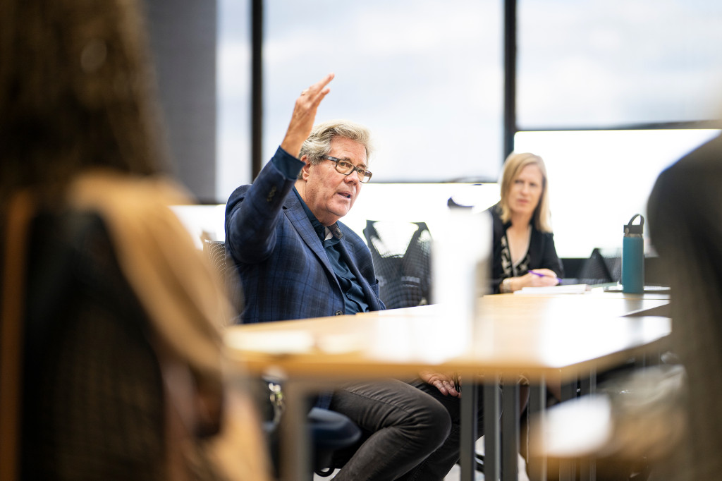 A man sitting at a table gestures with his hands and speaks. A student is visible in the background.