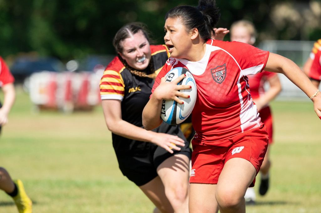 A woman in a red uniform runs hard with the rugby ball as an opponent clutches at her.