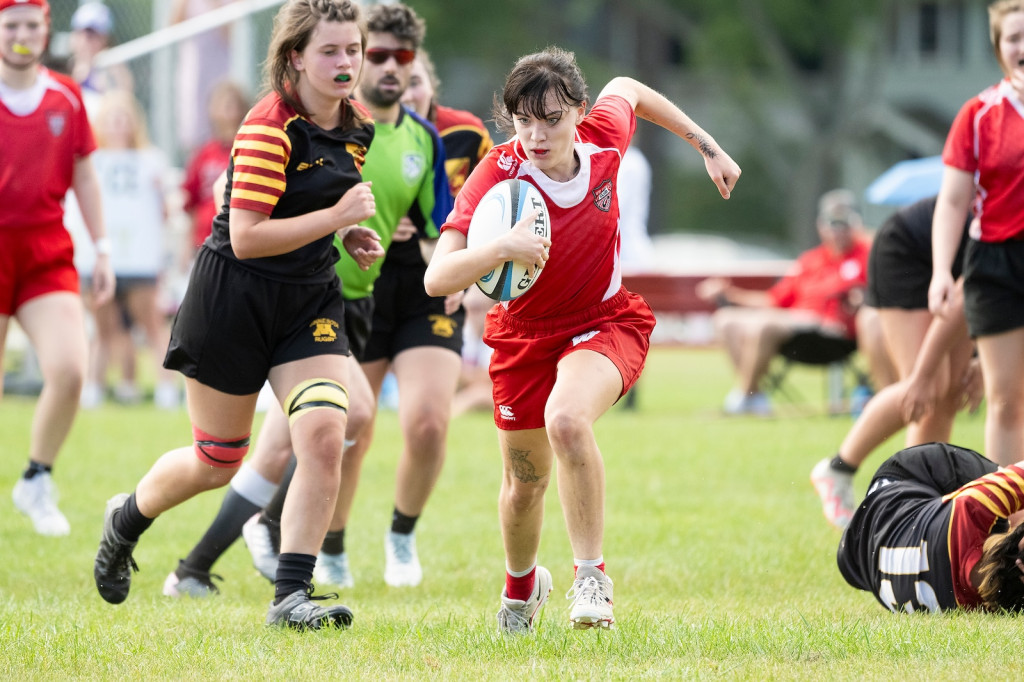 A woman holds on to the rugby ball and runs, knees high, as others pursue.