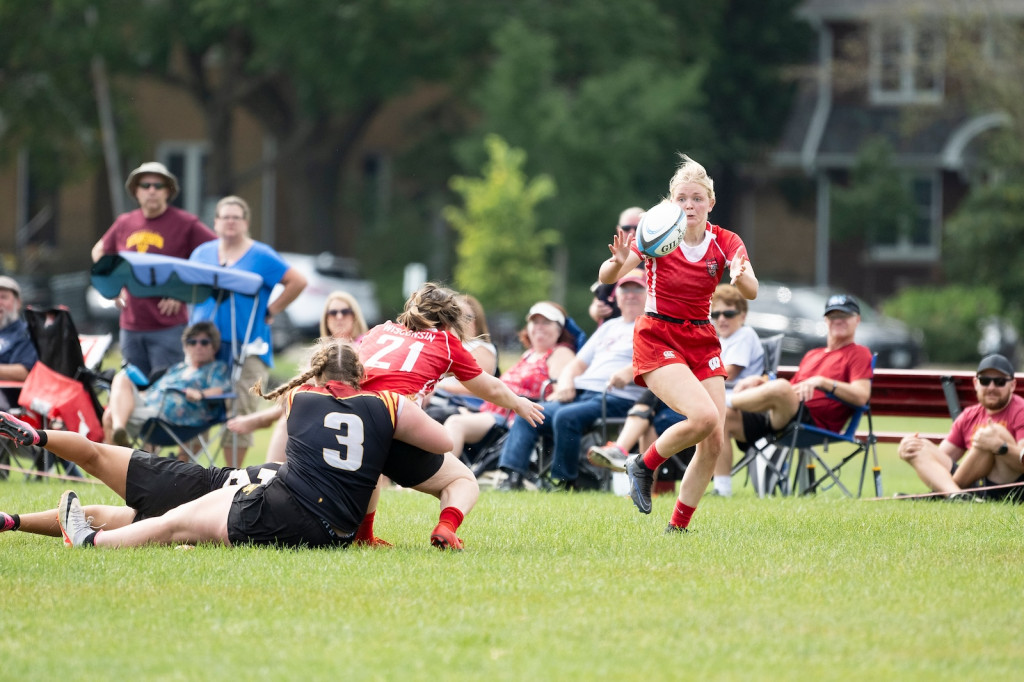 A woman being tackled tosses the rugby ball to another woman striding up the field.