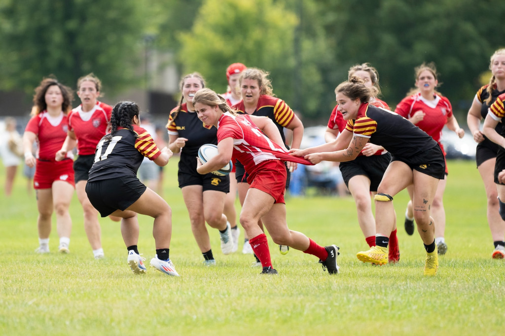 A woman in a red rugby uniform runs hard as another clutches the back of her jersey.