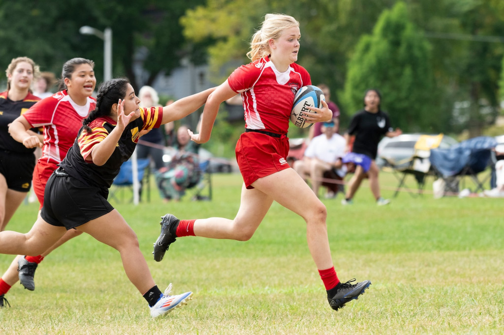 A woman in a red uniform runs with a rugby ball as one in a black and maroon uniform pursues her.