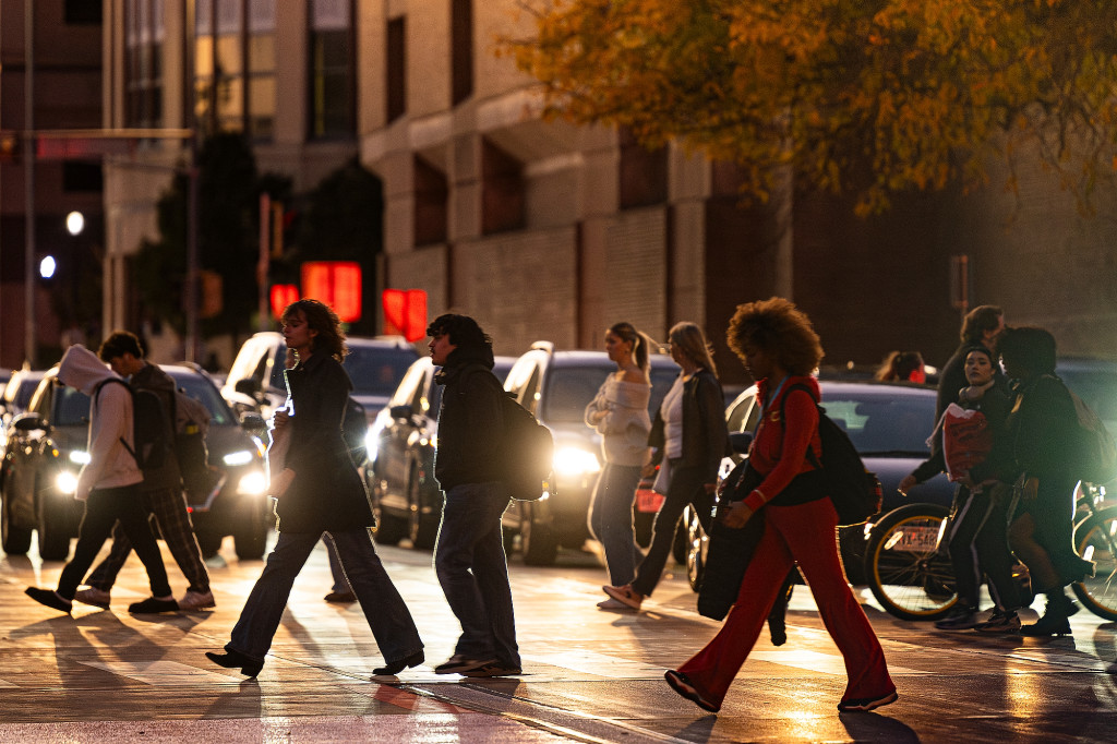 With the sun setting earlier in the day as the fall season sets in, pedestrians cross West Johnson Street illuminated by car headlights.