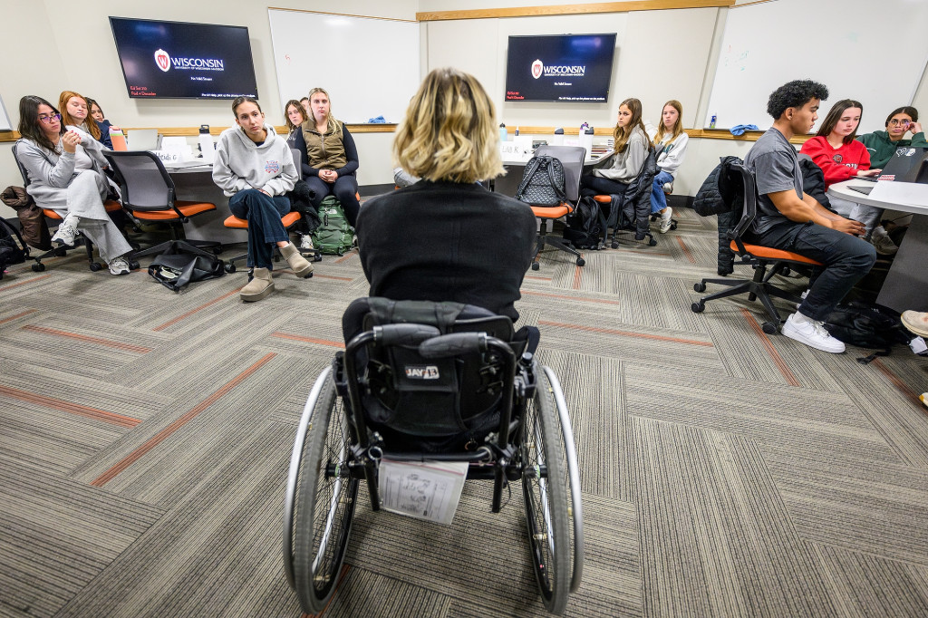 A view from behind as a woman in a wheelchair talks to a classroom.