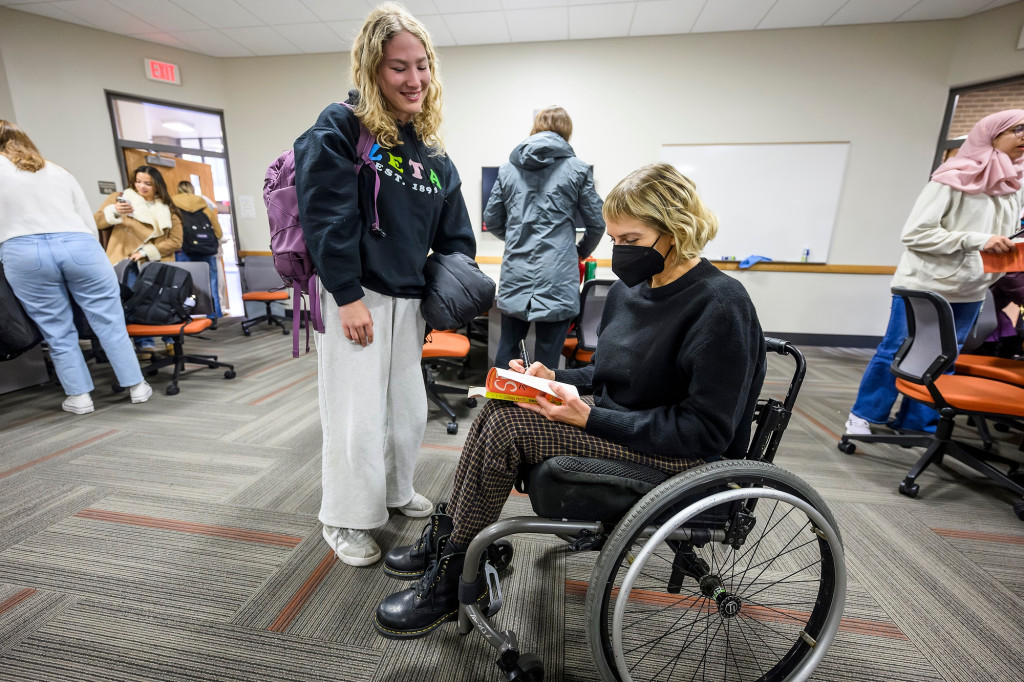 A woman stands next to another woman in a wheelchair, who's talking and signing a book.