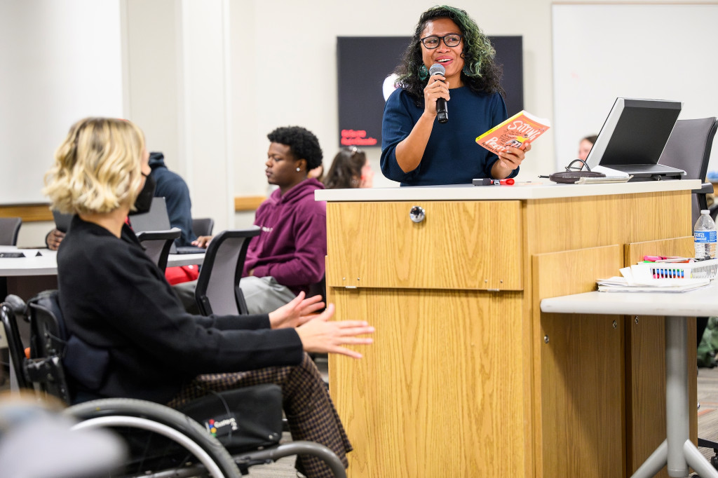 A woman at a podium holds a book and talks, while another in a wheelchair listens.