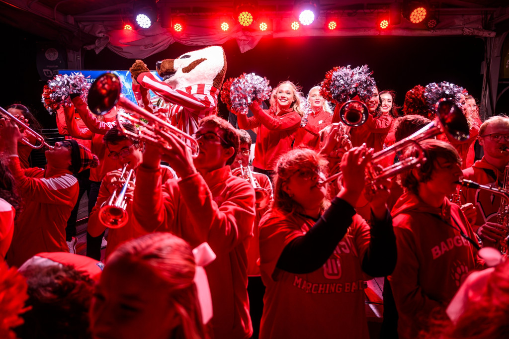 Band members and a person dressed in a Bucky Badger outfit cheer while bathed in red light.