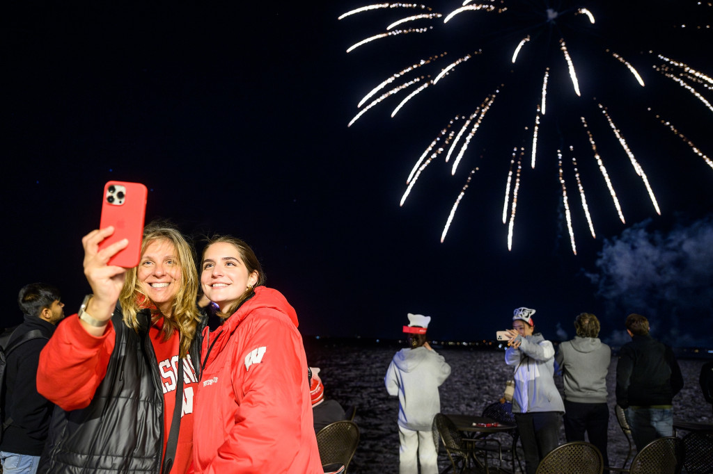 People pose for a selfie photo as fireworks ignite in the background.