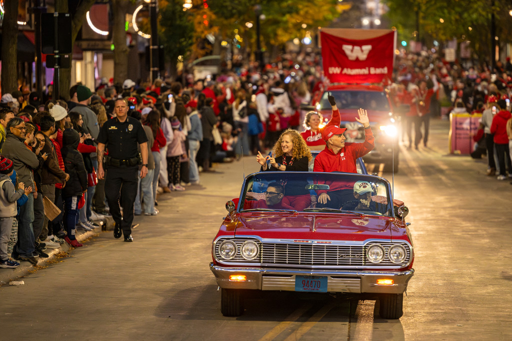 Two people sitting in the back of a convertible car wave to the crowd lining the street.