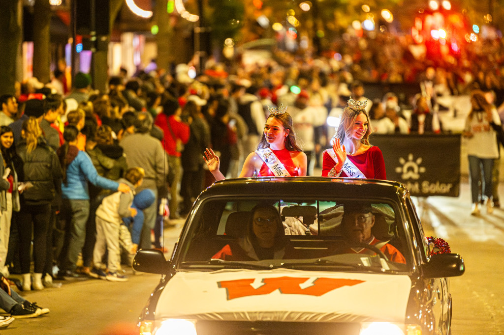 Two people in a convertible car wave to the crowd as the car drives down a street lined with spectators.