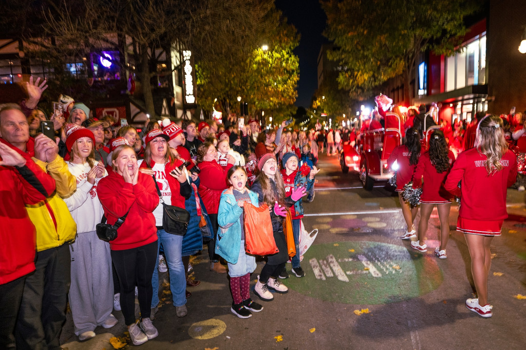 A group of excited children cheer as cheerleaders walk by. All are dressed in the red nad white of the University of Wisconsin.