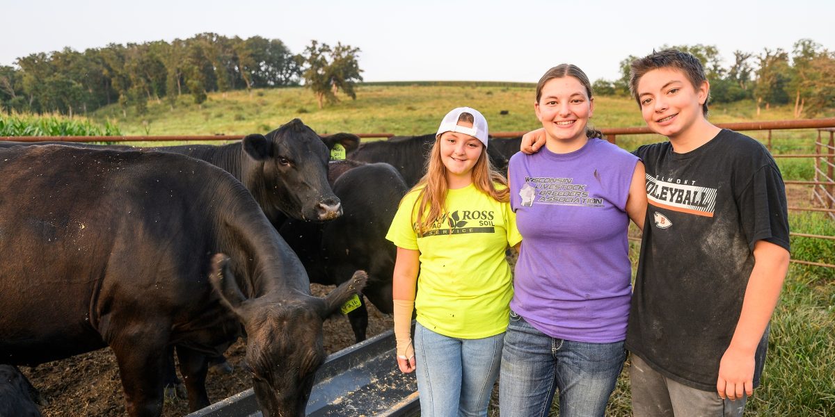 Three people wearing jeans and t shirts stand, arms around each other and smiling, as cows eat from a trough in the background.