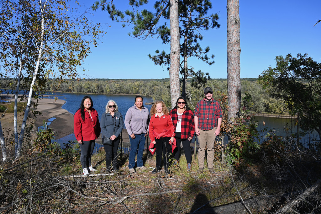 Six people stand on a wooded overlook of a lake and smile at the camera.