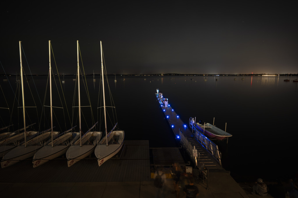 A nighttime view of a lake, with a pier jutting out into it, and colorful lights in the nighttime sky being reflected in the water.