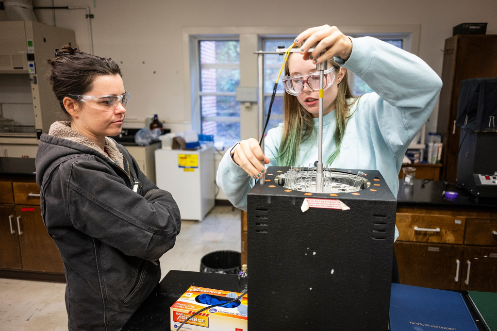 A woman works on lab equipment while another woman listens to her explain what she's doing.