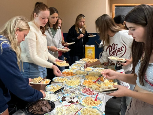 A group of people holding plates line both sides of a table filled with plates of cheese.