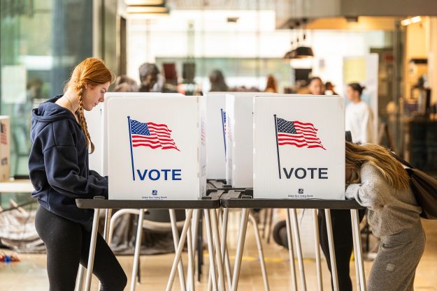 Two people stand filling out ballots at two voting booths across from each other.