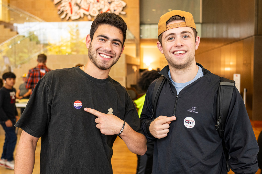 Two young man stand smiling and pointing to their stickers.