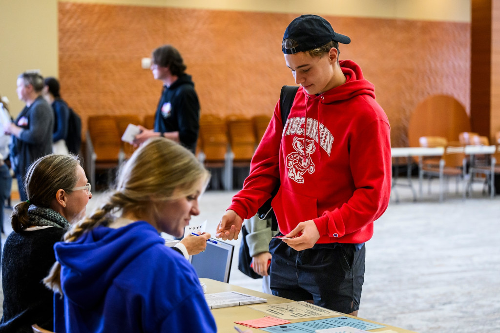 A standing man hands a ballot to a woman sitting at table.