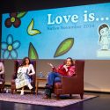 Two young women sit in chairs on a stage and listen while a third women, also in a chair, talks into a microphone.