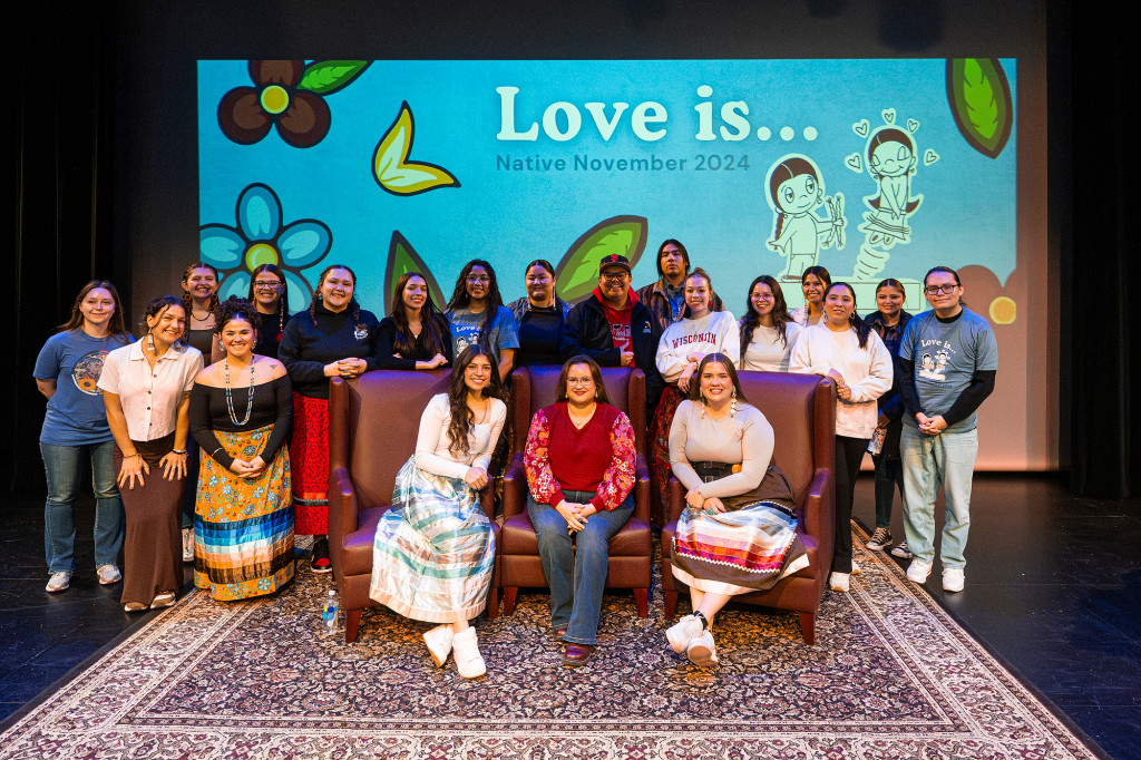 A group of people pose on a stage. Three sit in the front in chairs and the rest gather behind the chairs, standing.