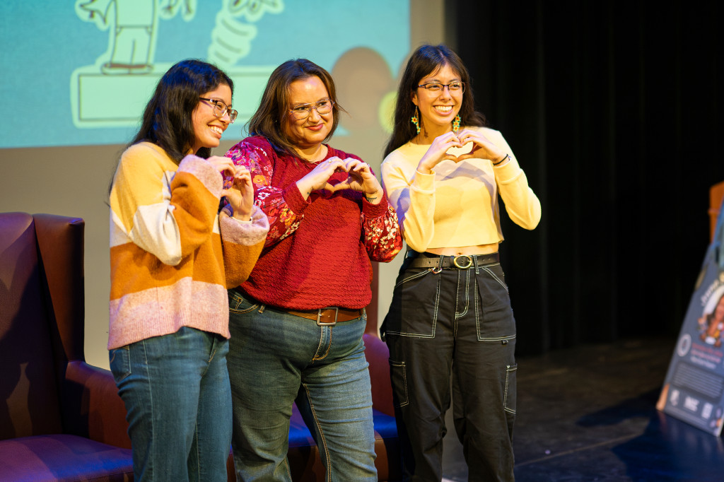 Three women pose for a photo. Each woman forms a heart with their hands and holds it over their chest.