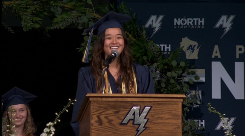 A woman in commencement robes speaks at a podium.