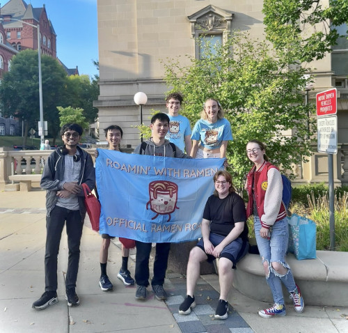 A group of people post for a photo on Library Mall; they're holding a flag that says "Roamin' with Ramen."