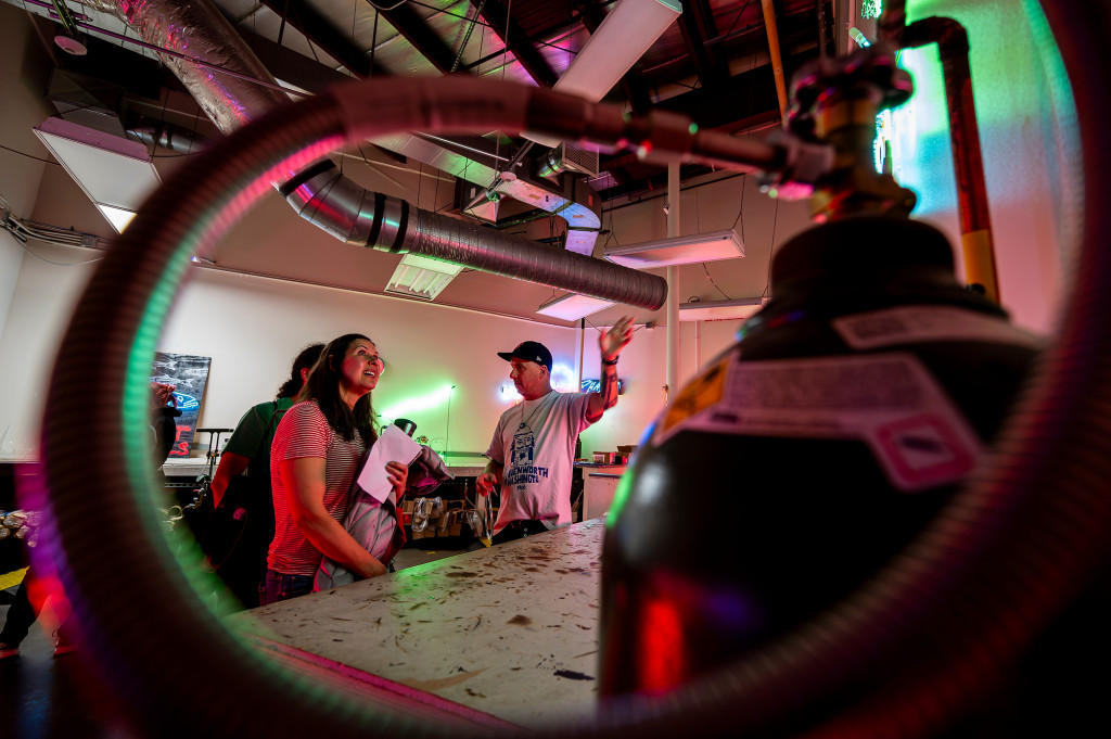 A man talks and gestures to several people, as they all stand in a room that has many neon lights in it.