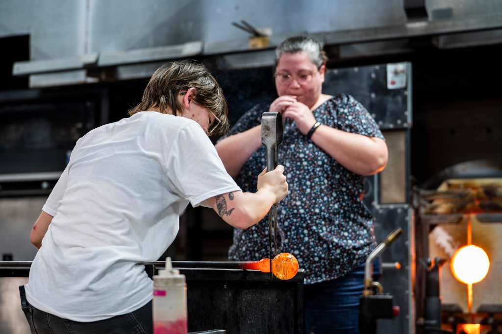 Two people work together; one blows on a pipe that's part of a glass-blowing device, the other tends to the orange bulb being created.