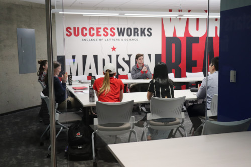 Several people are sitting around a table talking in a room with "Successworks" written on the wall.