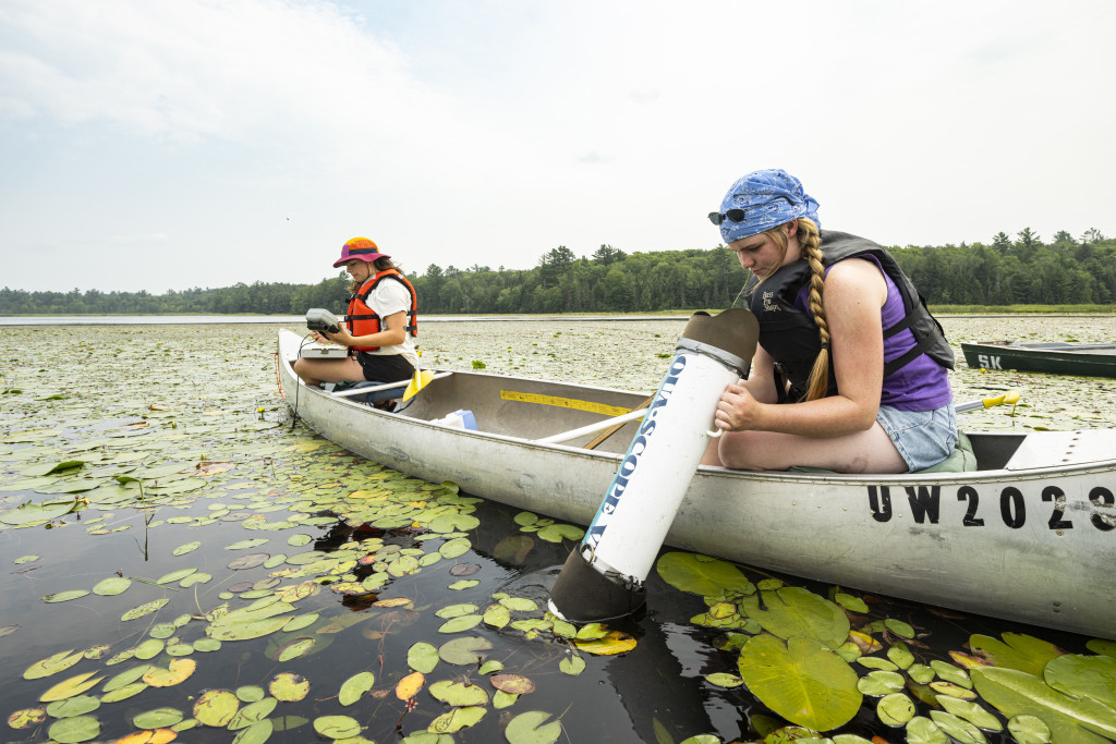 Two women in a canoe use equipment to take samples from the water.