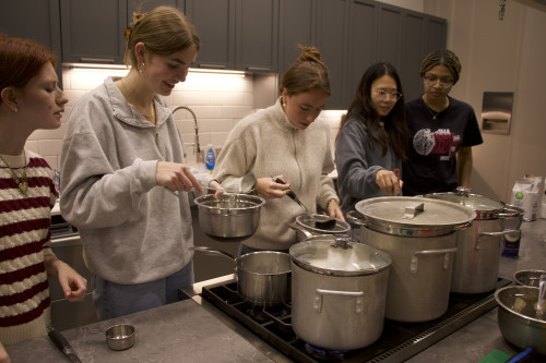 Four people line up in a kitchen in front of a stove where big pots of food are cooking.