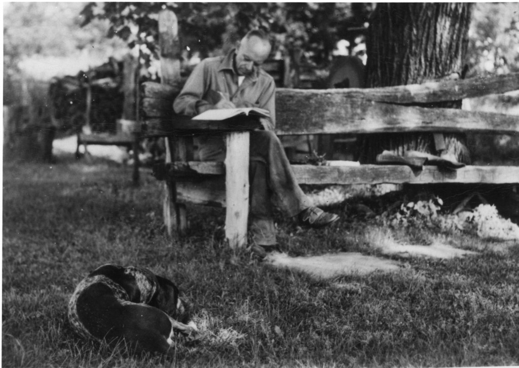 A man sits at a desk and writes, with a dog nearby.