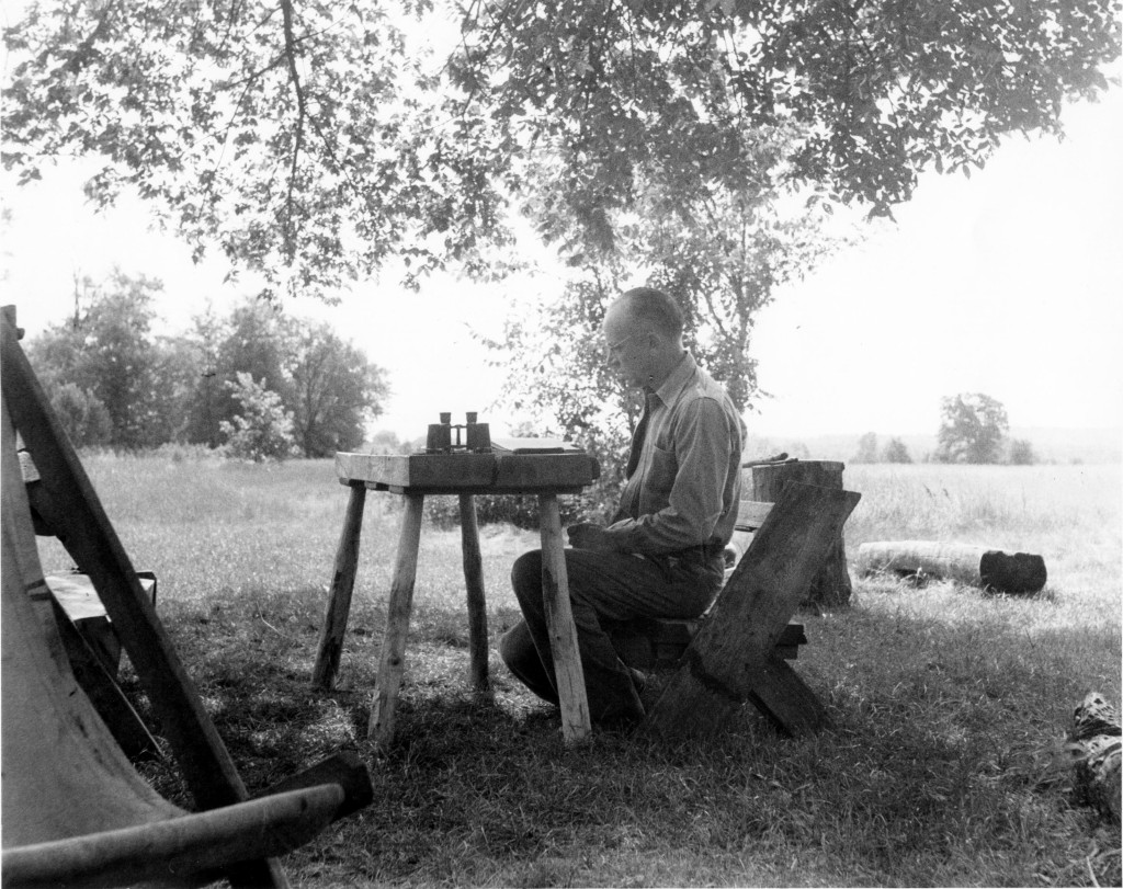 A black-and-white photo shows a man sitting at a table in an outdoors setting, writing in a notebook.