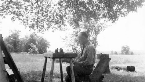 A black-and-white photo shows a man sitting at a table in an outdoors setting, writing in a notebook.