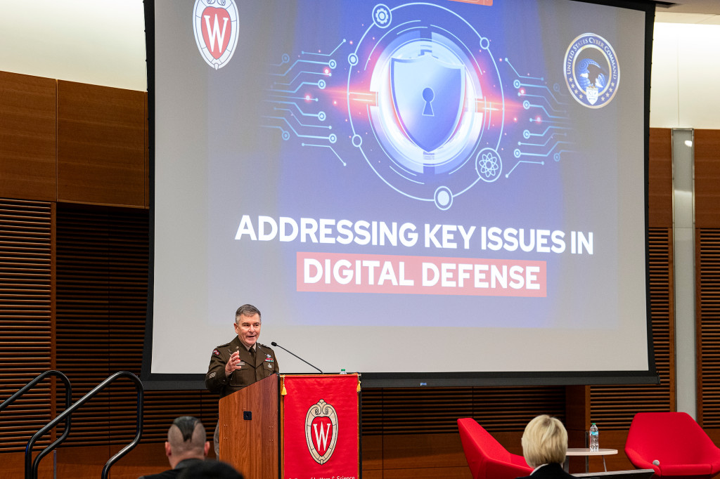 A man speaks at a podium in front of a large screen.