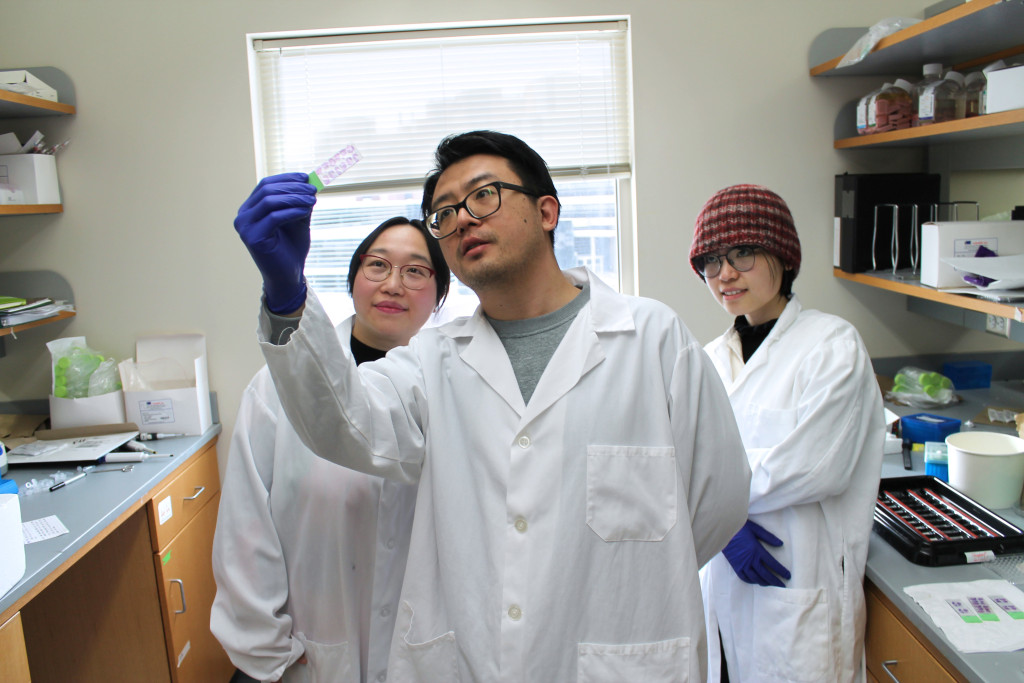 The scientists wearing white lab coats stand together looking at a sample that one of the scientist is holding up for examination. 