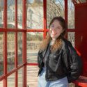 A woman is shown in a telephone booth painted red, with a building visible in the background.