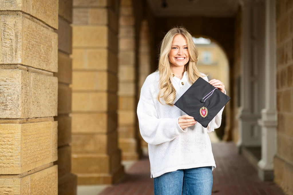 A woman in a white sweater holds a commencement cap with a University of Wisconsin logo on it.