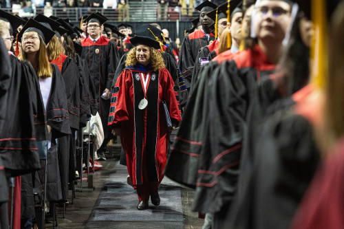 A woman in academic robes walks alongside graduates in their commencement garb toward the stage.