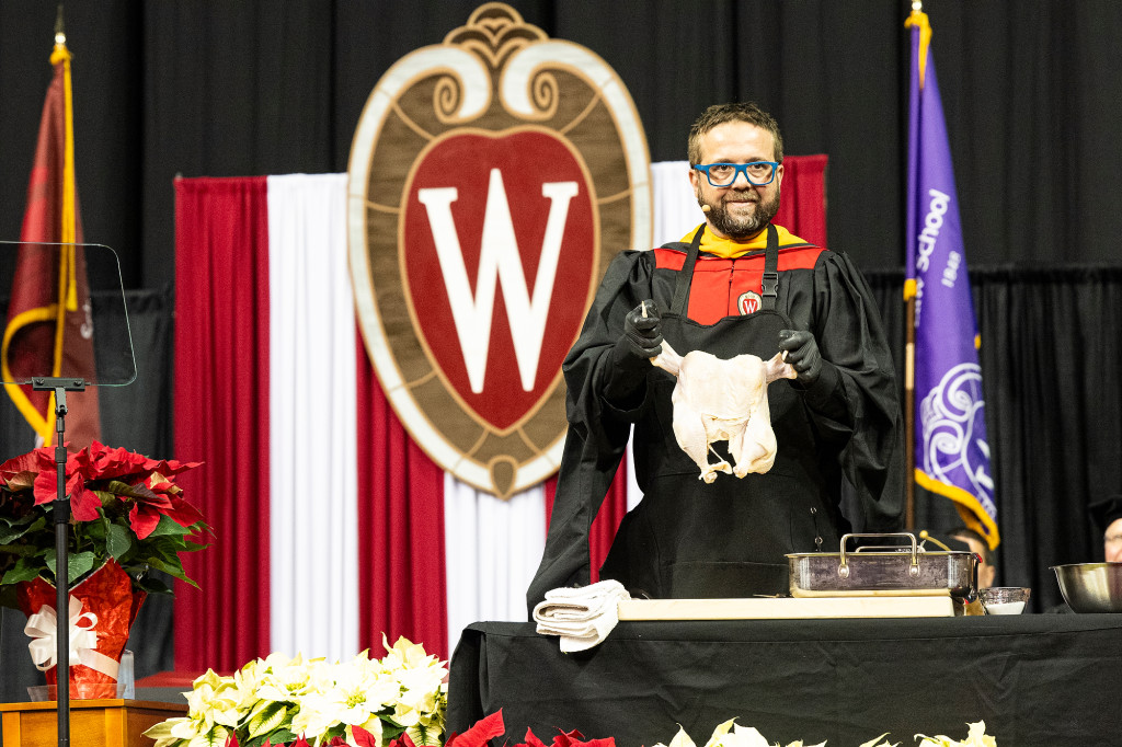 A man wearing academic robes on a stage holds up an uncooked chicken.