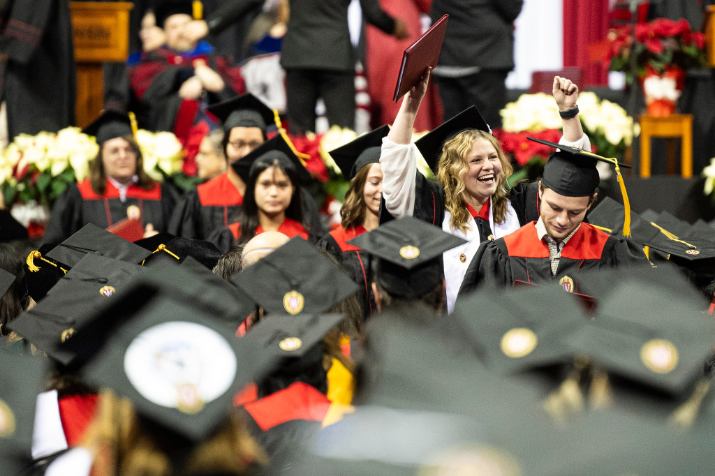 A group of people in black and red academic gowns smile and hold up diploma covers.