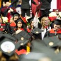 A group of people in black and red academic gowns smile and hold up diploma covers.