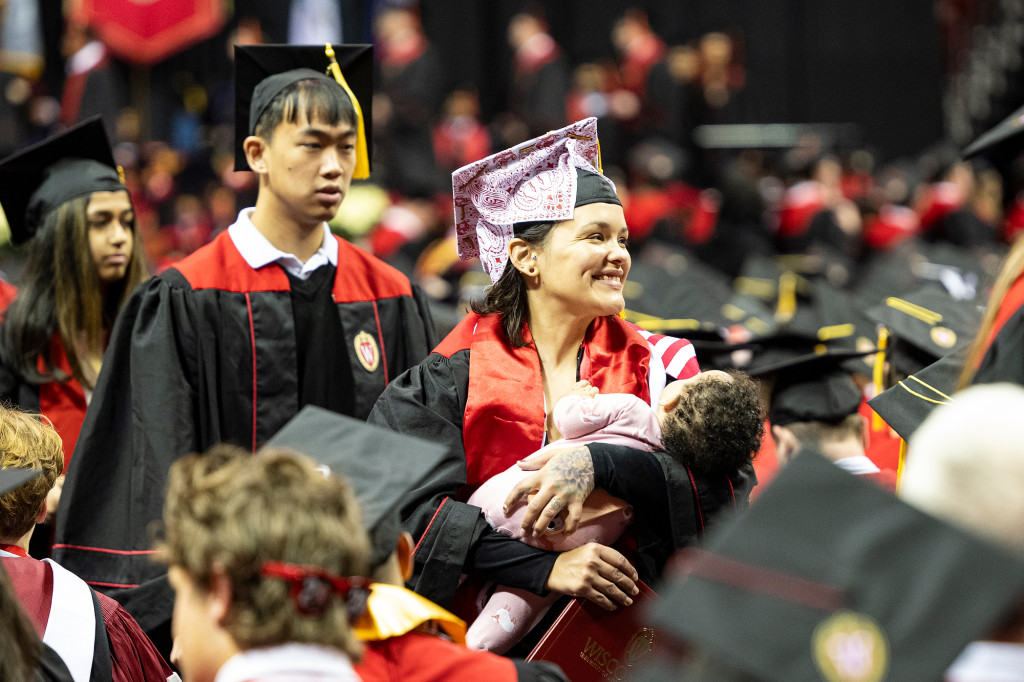 A woman in a graduation gown holds a baby and is surrounded by family members.