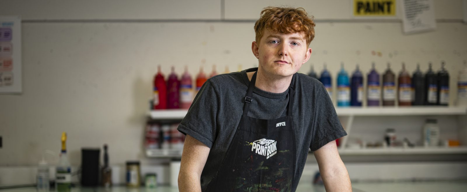 Photo of Bryce Dailey in a workshop apron in a screen printing studio as he leans with his hands on a work table.