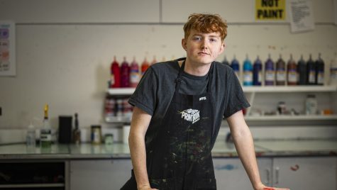 Photo of Bryce Dailey in a workshop apron in a screen printing studio as he leans with his hands on a work table.